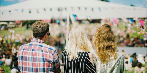 The backs of three people who look out over an outdoor event while the sun shines