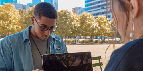 Two young people using electronic devices sit at a table outside in a park 