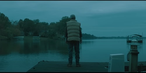 A man standing on a jetty, looking out over a still lake, with a Fincher-esque composition